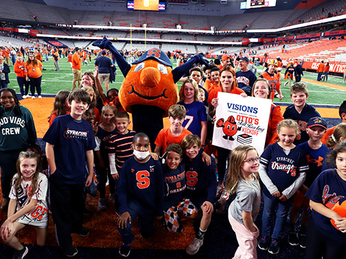 Lindsay (Visions Branding and Promotion Coordinator) and  Sarah (Community Development Liaison) pose with Otto the Orange and several students from local elementary schools on the SU football field for the Fall Reading Kickoff.