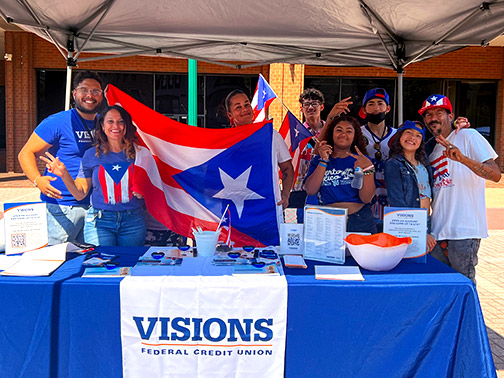 Community Development Liaison of Reading, PA, Gustavo, smiles in the sunshine at the Visions Table with several members sporting Puerto Rican flags, hats, and shirts.