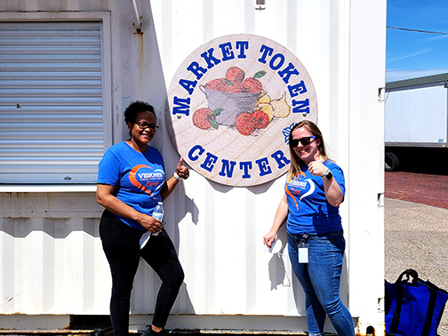 Visions employees Nadia and Sarah give a smile and thumbs up in front of the Market Token Center Sign at the Rochester Public Market.