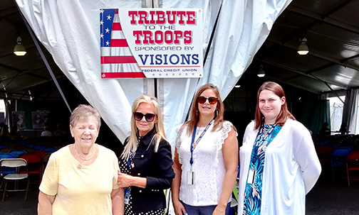 Photo is of Visions member, Ginny, and MCCEA Executive Board Members under the Visions Tribute to the Troops sign at the NJ State Fair.