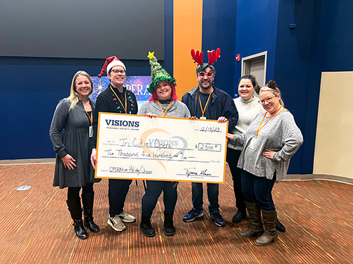 Dressed in festive holiday attire, Visions employee Jocelyn and members of the Tri-Cities Opera pose with a large check in the Auditorium located at Visions Headquarters.