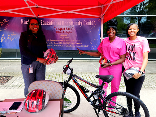 Regina (Visions FCU), Theresa Bowick (also known as “Nurse Bowick” and Founder of Conkey Cruisers), and DaSha Flowers (WDKX Intern) are pictured smiling in a small tent with a bicycle. They are holding helmets and proudly pointing at a banner that says “Rochester Educational Opportunity Center”.