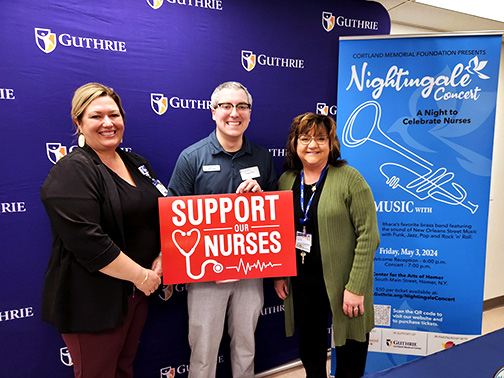 Kansas Underwood (VP of Nursing of Guthrie Cortland Medical Center), Mark (Visions FCU Branch Manager), and Shelia Ossit (Director of the Cortland Memorial Foundation) are smiling for the camera in front of a purple wall that contains the Guthrie logo and a pop-up sign about the Nightingale Concert. Mark is holding a sign that says “Support our nurses”.