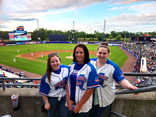 Visions representatives Sarah, Kenda, and Jessie smiling in front of the Syracuse Mets baseball field on a beautiful spring evening. They are wearing the Syracuse Mets star-spangled jerseys.